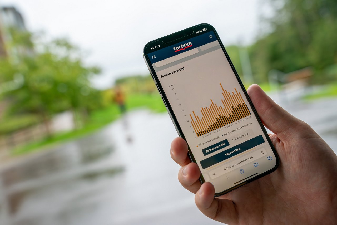 Personen holder en iPhone, og skjermen viser Techem Beboer App. Grafen på skjermen illustrerer forbruksoversikten.