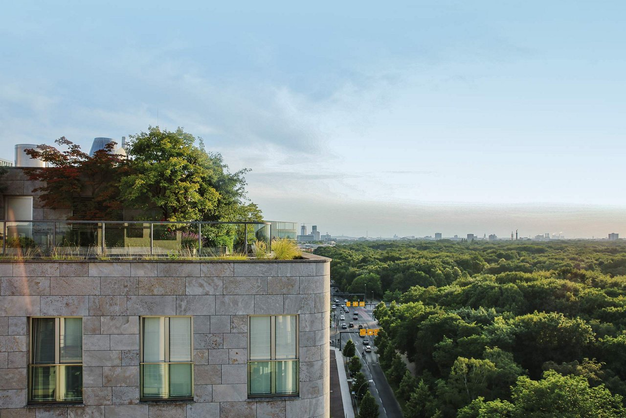 Eine Dachterrasse eines modernen Gebäudes mit einer begrünten Fläche. Die Terrasse ist mit Bäumen und Pflanzen bepflanzt und bietet einen Blick auf eine weitläufige, grüne Landschaft mit einem Wald und einer Straße mit Autos im Hintergrund.