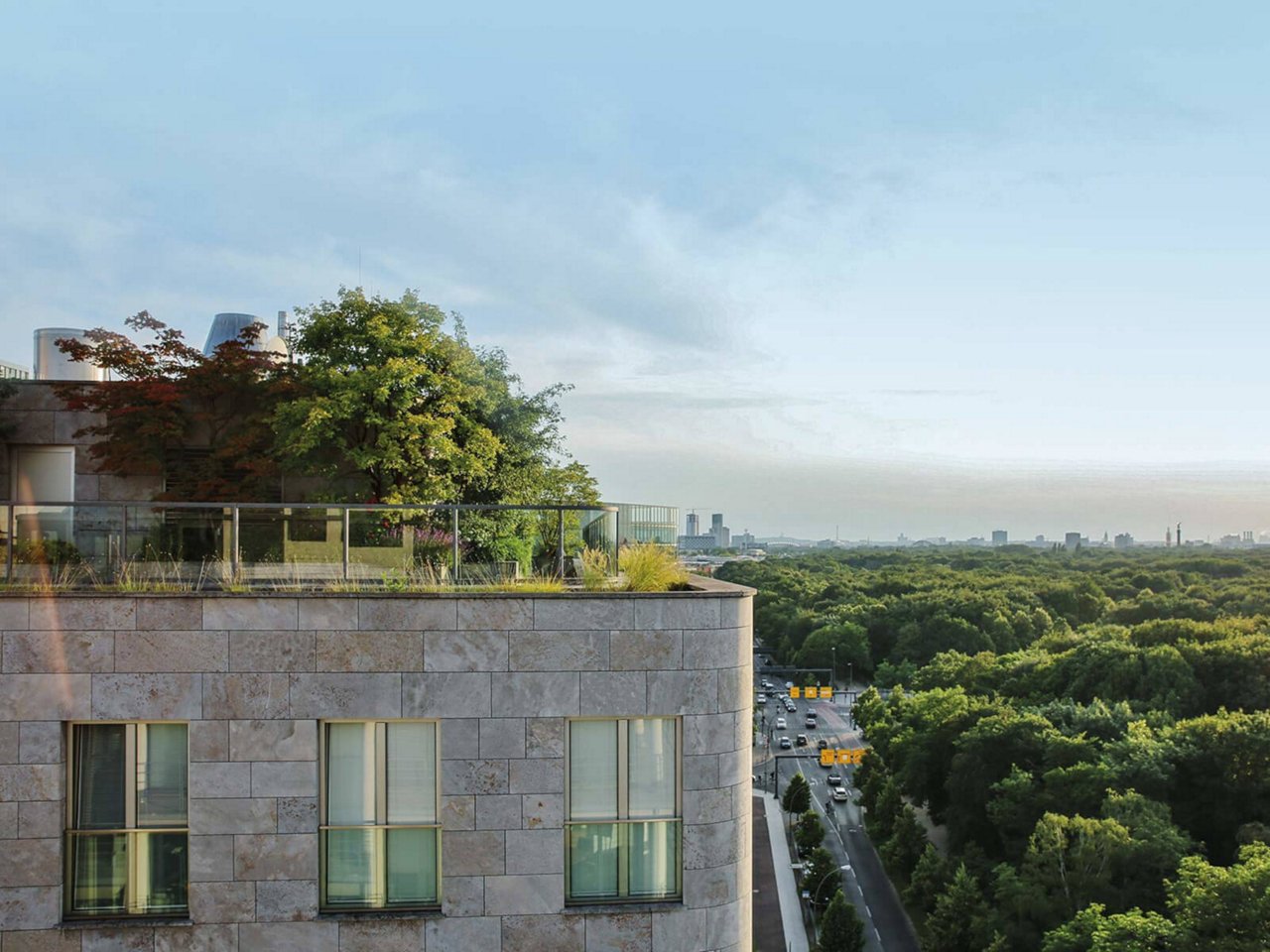 eine Dachterrasse eines modernen Gebäudes mit einer begrünten Fläche. Die Terrasse ist mit Bäumen und Pflanzen bepflanzt und bietet einen Blick auf eine weitläufige, grüne Landschaft mit einem Wald und einer Straße mit Autos im Hintergrund.