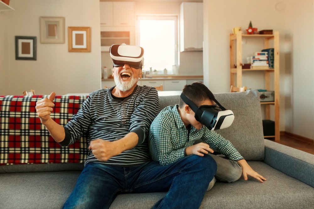 Begeisterter älterer Mann mit VR-Brille auf dem Sofa zu Hause mit lachendem Enkel neben ihm