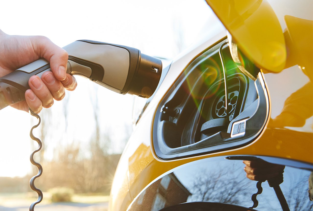 Eine Männerhand hält ein Ladekabel an ein gelbes Elektroauto. Das Laden findet im Freien bei Tageslicht statt, was auf umweltfreundliche Mobilität hinweist.