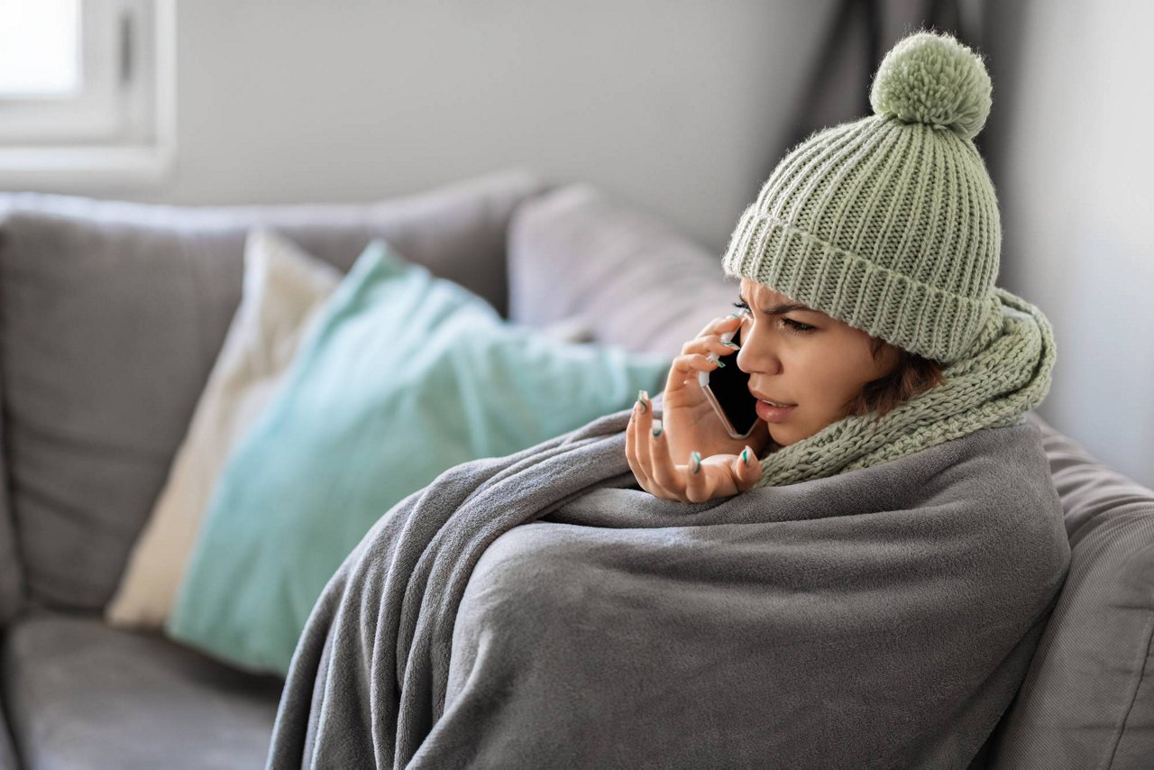 Stressed Young Woman Talking On Cellphone While Sitting On Couch Wrapped In Blanket, Upset Female Tenant Freezing At Home, Complaining To Landlord About Low Temperature In Room, Side View; Shutterstock ID 2231336657; purchase_order: TES 0000; job: Motiv für Kundenpräsentation Digitaler Heizungskeller; client: Techem/faust&auge; other: 