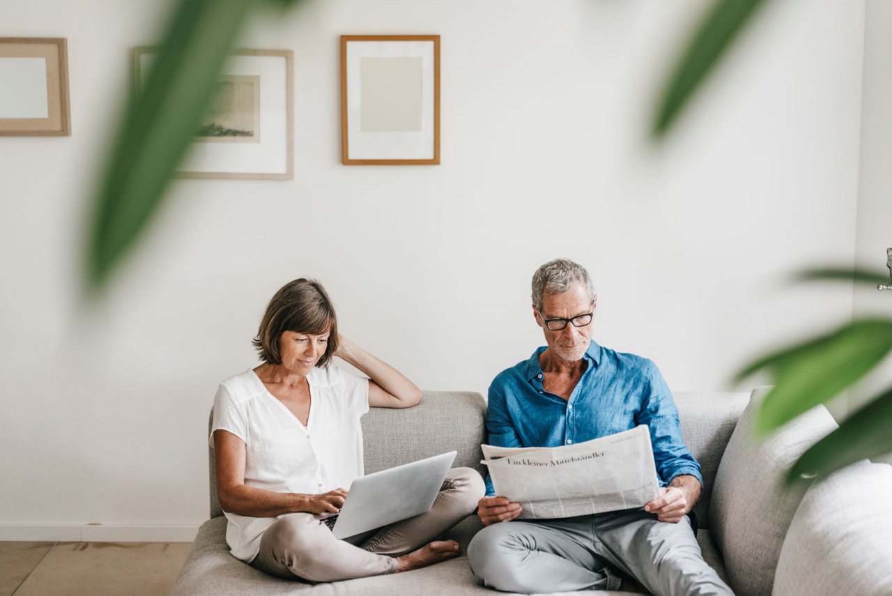 En kvinne og en mann som sitter komfortabelt i en sofa. Kvinnen har en laptop i fanget, og hun ser konsentrert ut mens hun arbeider eller surfer på nettet. Mannen ved siden av henne leser en papiravis.