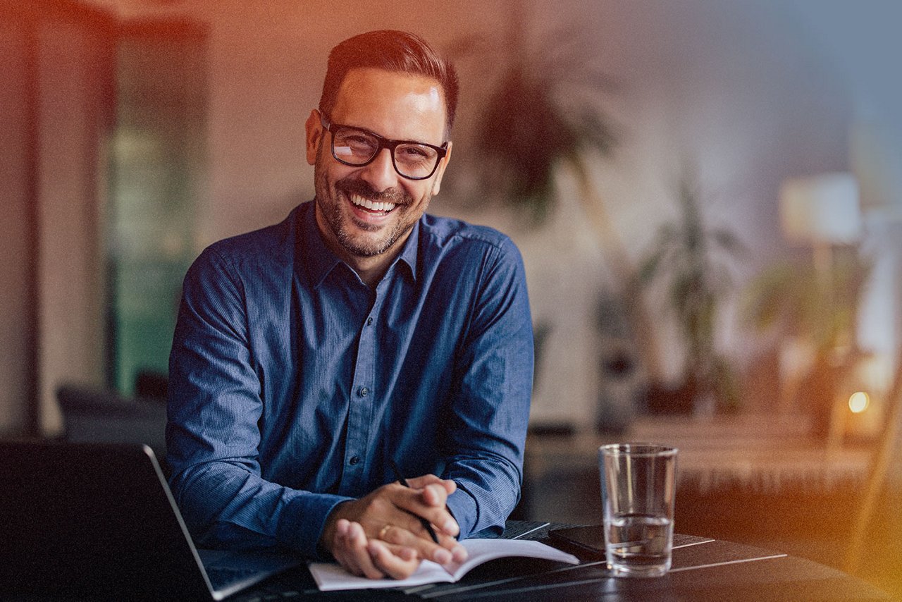 Ein Mann mit Brille in einem blauen Hemd sitzt an einem Bürotisch und lächelt in die Kamera.