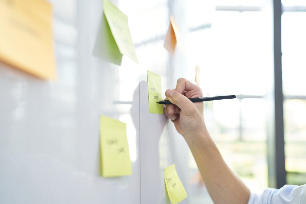  En hånd som strekker seg mot en tavle i et møterom. På tavlen er det festet flere post-it lapper i forskjellige farger, som gir et livlig og kreativt inntrykk.