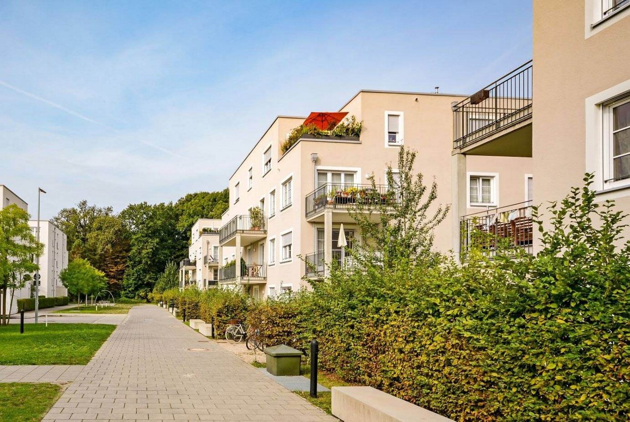 Das Bild zeigt eine moderne Wohnsiedlung mit hellen, mehrstöckigen Gebäuden, die über Balkone und viel Grün in Form von Hecken und Bäumen verfügen. Ein gepflasterter Gehweg führt durch die Anlage, die eine ruhige und gepflegte Atmosphäre vermittelt, umgeben von Natur und nahe an einem Waldgebiet.
