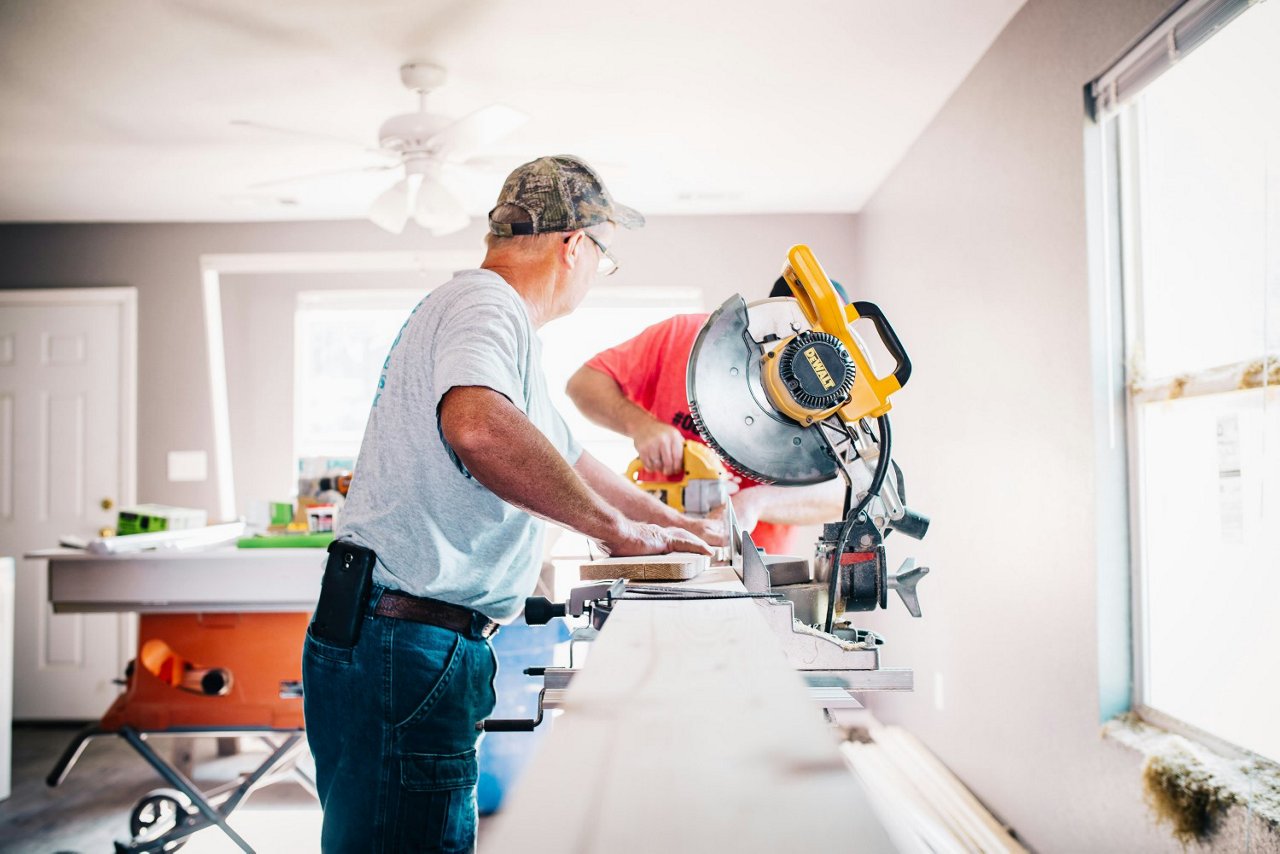 Zwei Handwerker schneiden Holz in einem renovierungsbedürftigen Raum. Einer bedient eine Kappsäge, der andere arbeitet mit einem Elektrowerkzeug. Baustellenmaterial liegt im Hintergrund