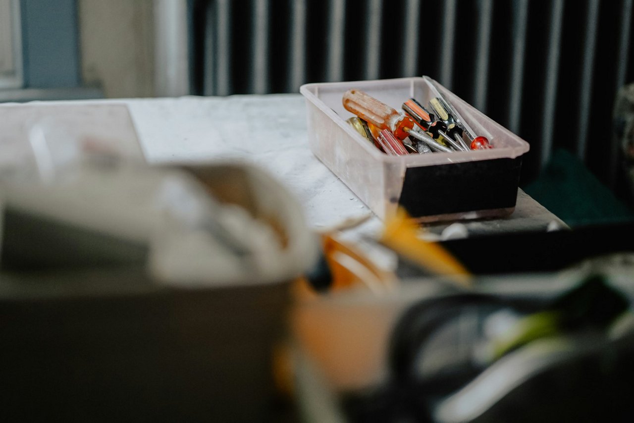 Ein transparentes Kunststoffbehältnis mit verschiedenen Schraubenziehern liegt auf einer Werkbank. Im Vordergrund sind unscharfe Werkzeuge und Materialien zu sehen.