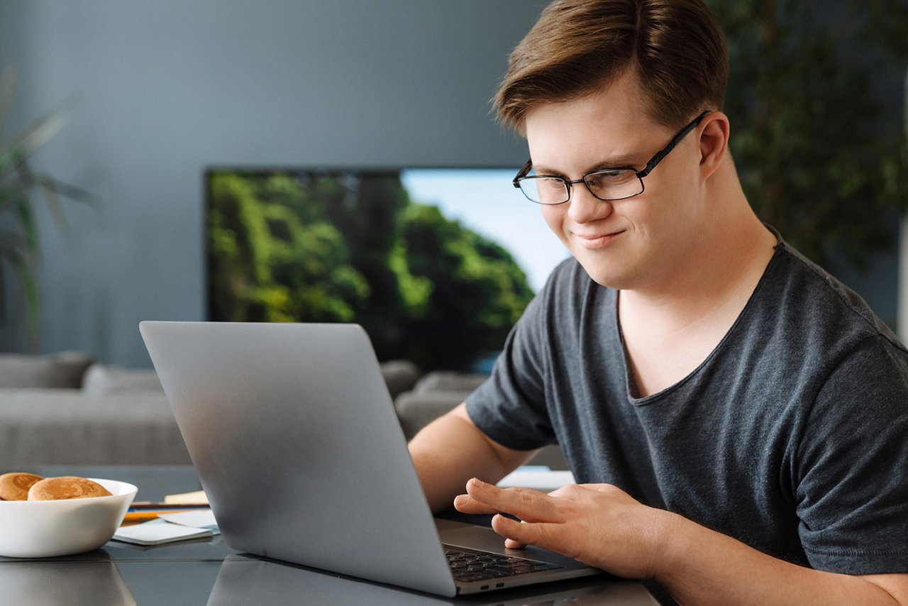 Junger Mann mit Down-Syndrom und Brille arbeitet lächelnd an einem Laptop am Tisch