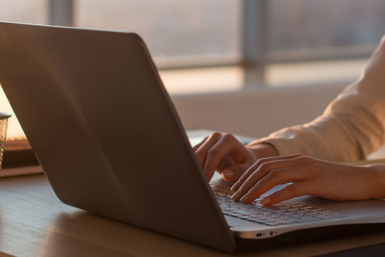Eine Person, die an einem Laptop arbeitet. Die Hände der Person sind auf der Tastatur zu sehen, während der Laptop auf einem Holztisch steht. Der Hintergrund ist unscharf und zeigt ein Fenster mit Sonnenlicht. Der Fokus liegt auf den Händen und dem Laptop.