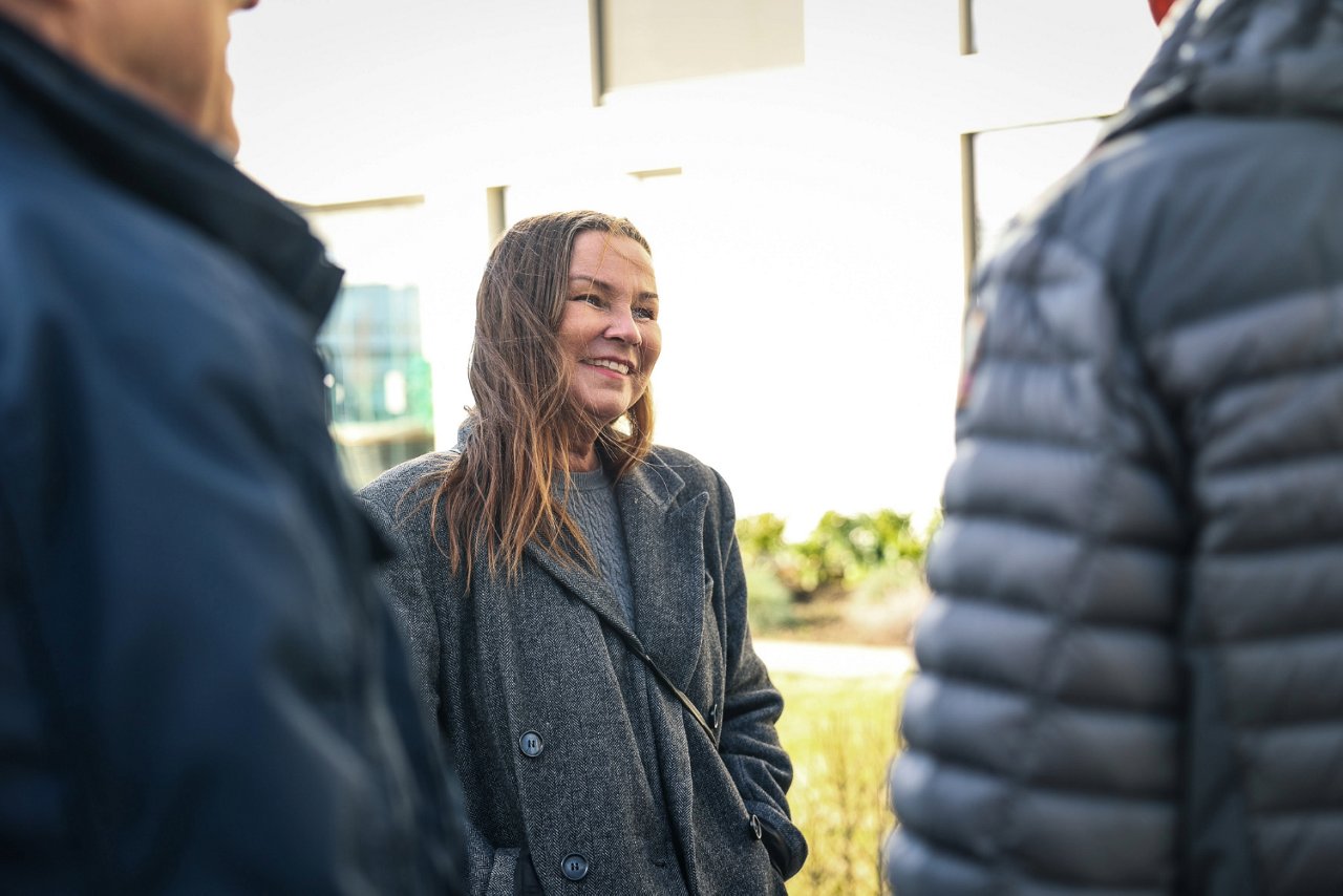 Bildet viser en dame som smiler og har hendene i lommene. Hun ser ut til å være i en samtale med to personer, hvor vi ser ryggen deres.