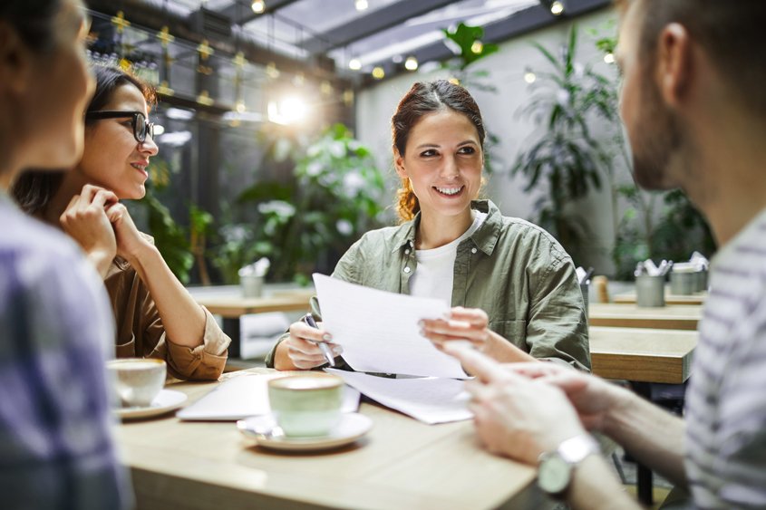 Kolleg*innen sitzen gemeinsam am Tisch, trinken Kaffee und besprechen Dokumente