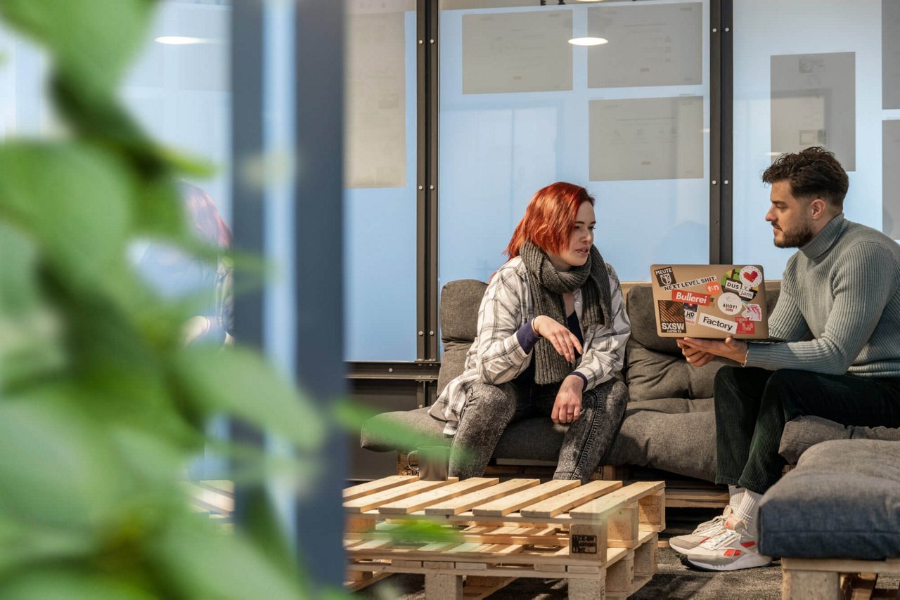 Eine Frau und ein Mann sitzen gemeinsam auf einem Sofa. Der Mann hat einen Laptop in der Hand und scheint der Frau etwas zu erklären. Vor ihnen steht ein Palettentisch. 
