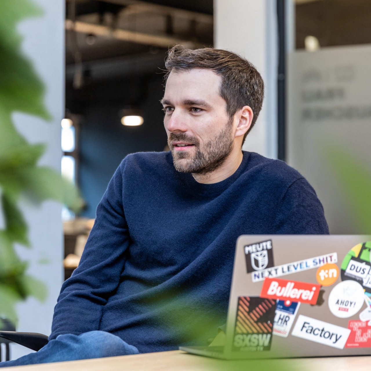 Ein Mitarbeiter mit brünetten, kurzen Haaren und einem dunkelblauen Pullover sitzt am Laptop und schaut zur Seite
