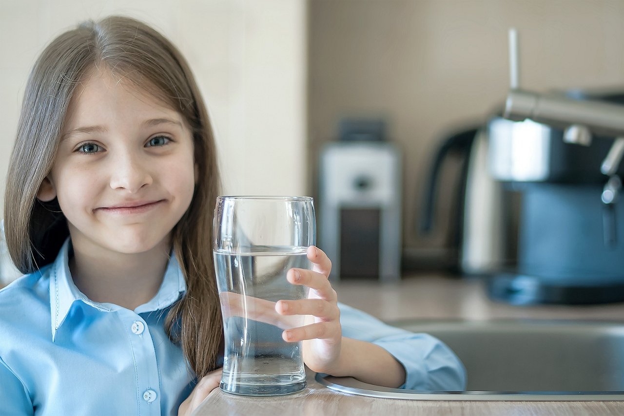 Kind mit langem Haar in blauem Hemd hält ein großes Glas mit Trinkwasser in der Hand