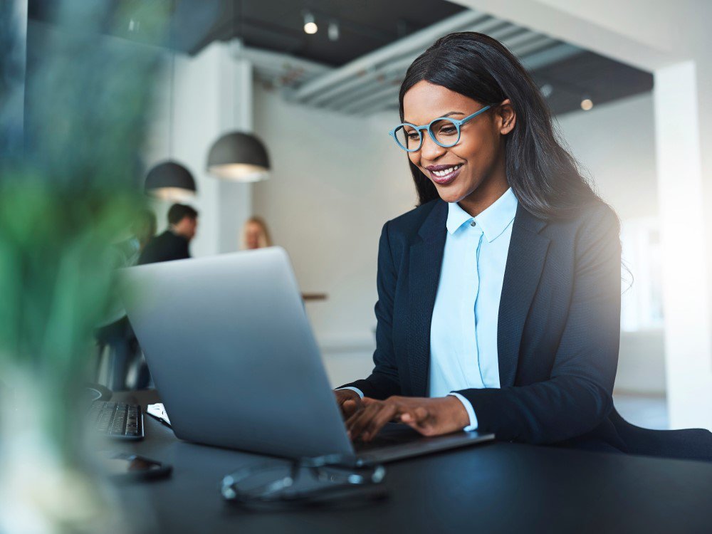 Eine junge Frau mit einer hellblauen Brille sitzt im Büro vor einem Laptop und lächelt. Im Hintergrund sieht man weitere zwei Kollegen.